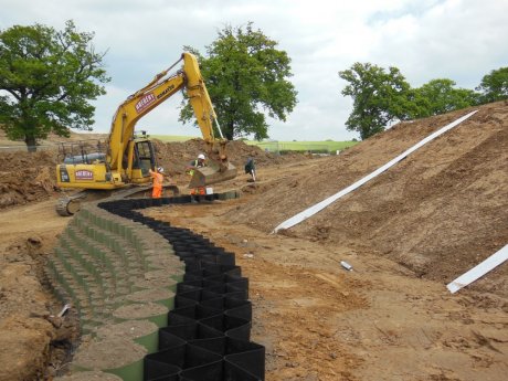 The design incorporated a Webwall front face, Trigrid geogrid reinforcement and Fildrain drainage geocomposite. Here the front green cells are being filled with topsoil. Fildrain structural drainage strips are visible on the cut slope
